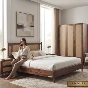 A woman sits on a wooden bed in a styled bedroom featuring a matching wooden wardrobe with cane doors, bedside tables, lamps, and a neutral-toned rug. The brand name Wooden Empiro FURNITURE appears in the bottom right corner.