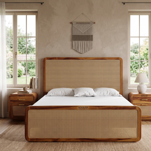 A wooden bed frame with a woven cane headboard and footboard, styled in a bright, modern bedroom with two windows, matching nightstands, and a textured wall hanging.